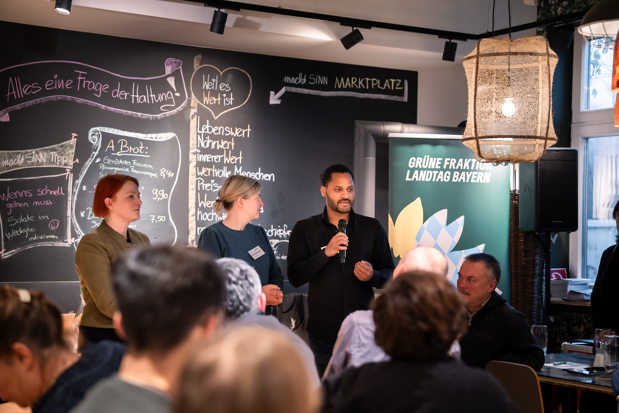 Mia Goller, Katharina Schulze und Benjamin Adjei stehen vor Publikum und sprechen in ein Mikrofon.